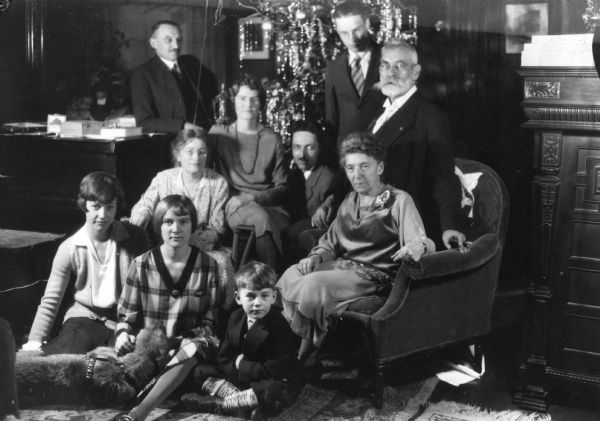Group portrait of members of the family of Ferdinand Hotz posed in front of the Christmas tree in the Hotz home. They are, standing in the back, left to right: Ferdinand Hotz, son Ferdinand Leonard (Fedy) Hotz, and cousin Rudolph Schenck. Middle row, left to right, Ferdinand's wife Clothlide Schmidt Hotz, daughter Alice Hotz Apfelbach, son-in-law Rudi Schmid (married to the photographer's daughter Helen, not pictured), and cousin by marriage, Frieda Bohm Schenck. Front row, left to right, Clothilde's cousin Elsie Wieczorowski, daughter Margaret (Sissy) Hotz, and Henry W. Apfelbach, Alices' step son. Sissy cradles a dog's head on her lap.<p>Rudolph Schenck's mother, Elizabeth Hotz Schenck was the sister of Ferdinand Hotz's father Phillip.
