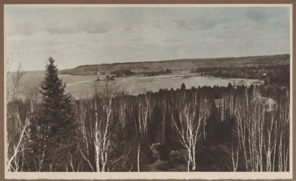 Elevated view, looking north, from the Hotz family compound near the bluff entrance to Fish Creek. Evergreens and bare birch trees stand in the foreground, with Fish Creek Harbor and Hen Island beyond. On the right is the school house; in the background are the bluffs of Peninsula State Park, largely devoid of trees due to logging.