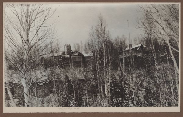 View from below a bluff, through evergreens and birch trees, looking up at the Hotz Fish Creek compound. The stone garage and tower are on the left.