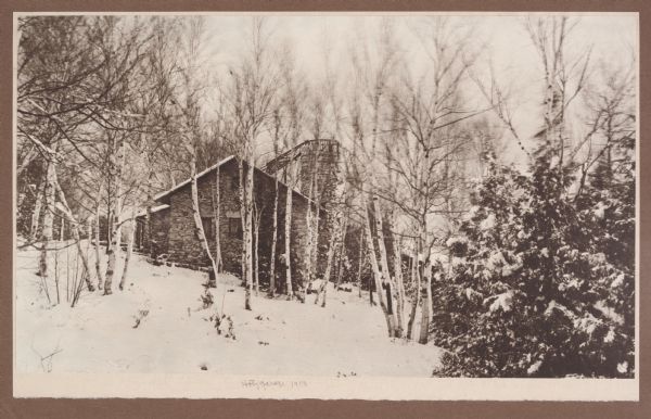 View of the side of the stone garage and tower at the Hotz family compound at Fish Creek. There is a rustic ladder from the garage roof to the observation deck at the top of the tower. Snow covers the ground and clings to birch and evergreen trees.