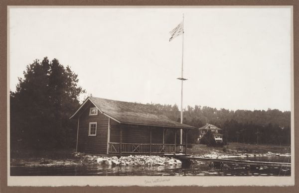 View from water of simple frame structure with rustic porch rail standing on the Fish Creek beach. A pier extends from the porch and a flag flies on a tall flag pole at the side of the bathhouse. There is a two-story, foursquare style house in the background, identified as the Vorous house.