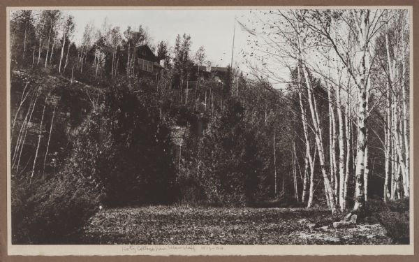 View from the base of the bluff looking up at the Hotz compound. A flag blows in the breeze above birch and evergreen trees. A woman is standing on the porch of the building on the right.
