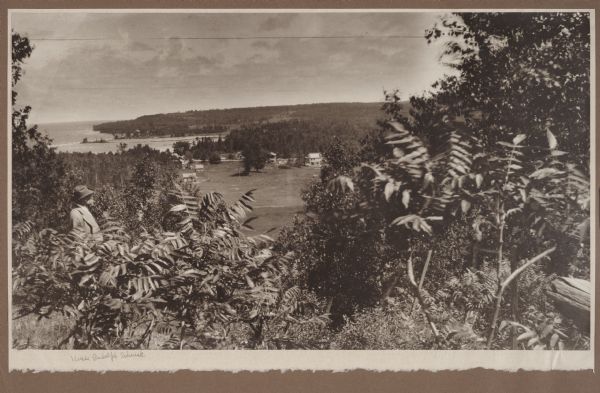 A well-dressed man identified as Uncle Rudolph Schenck takes in the view from a bluff overlooking the Fish Creek valley and harbor. There are houses and outbuildings in the middle distance; the bluffs of Peninsula State Park are in the background.