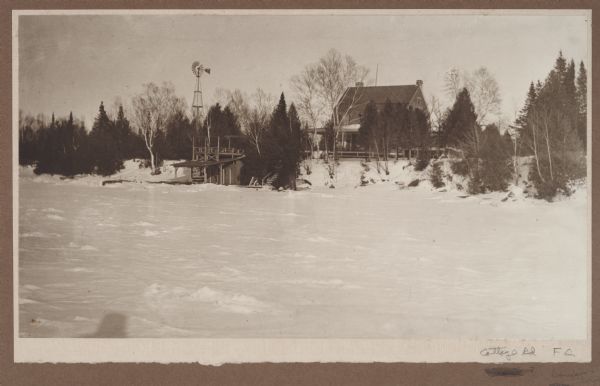 Winter view, taken from the frozen Green Bay, of a house south of Fish Creek. There is a windmill in the yard. A boathouse with upper deck and railing stands on the shore. Steps alongside the boathouse lead from the yard to the shore.