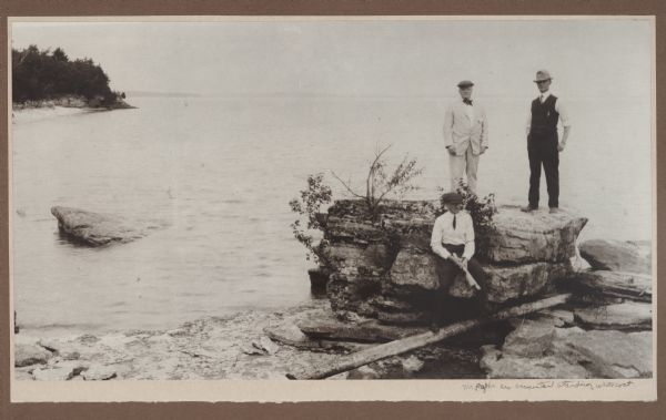 Mr. Paepcke, identified as an accountant, in white suit, poses with two unidentified men on large rocks on the shore of Europe Bay. Plum Island and Detroit Island are in the background.