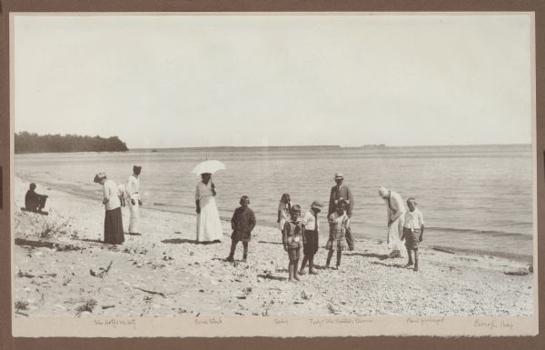 A group of children and adults enjoy a sunny day at the beach. Those identified include Mrs. Ferdinand (Clothilde Schmidt) Hotz, on left with hat and light jacket, and her husband with face turned toward the camera; Erna Steck, with parasol; Margaret (Sissy) Hotz, in white dress near the water; Ferdinand Leonard (Fedy) Hotz, with necktie and hat; Mr. Albert Trostel in hat and suit with his daughter Elinor in front of him; and at far right, Paul Guenzel. The boy next to Fedy with a large neckerchief is Albert Trostel, Jr. Plum and Detroit Islands are visible in the background.