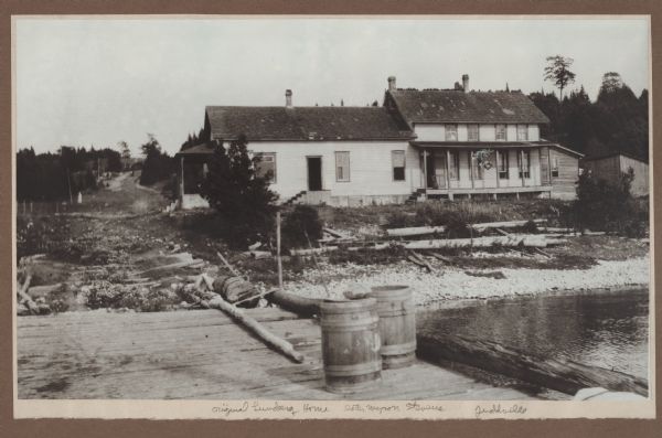 An older story and a half wood frame house with a one-story addition on the left and lean-to on the right stands near the Green Bay shore. It is identified as the "original Lundberg home, later Myron Stevens." Two barrels sit on a dock in the foreground; Juddville road extends up the hill from the dock.