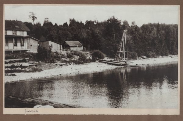 View from pier of the Lundberg home, with outbuildings along the Green Bay shore at Juddville. A platform with tower and ladder rests on the beach. This likely is a gin pole (or pole derrick), used in stepping the mast on a sailboat or iceboat. A log portion of the Juddville dock is seen at lower left.