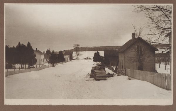 Winter scene from Egg Harbor Road (Highway 42) in Fish Creek looking north toward frozen Fish Creek Harbor with the bluffs in Peninsula State Park beyond. Evergreens and houses line the snow-covered road.  Cordwood is stacked on the left. There is a horse-drawn sleigh on the frozen bay.