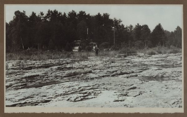 View across rocky shoreline of Dr. Gordon standing in front of his cottage on the rocky North Bay shore. The cottage is partially obscured in a dense stand of evergreen trees.