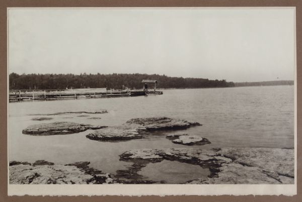 View from rocky shoreline of a man with a fishing pole standing in the shade provided by a small roof at the end of Dr. Gordon's pier. Fishing nets dry along the railing.