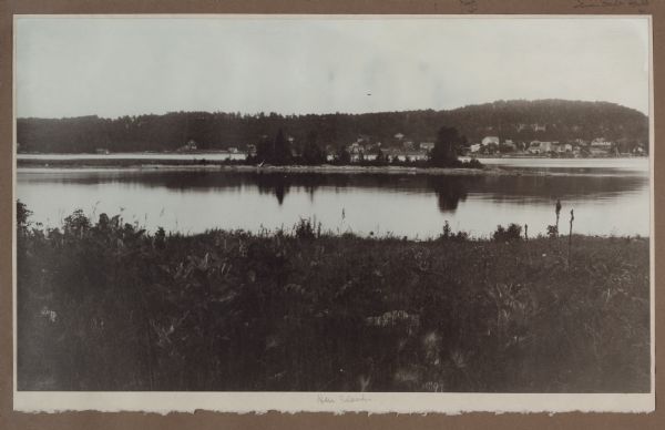 Hen Island is reflected in the water of Fish Creek Bay in this view from Nelson Point in Peninsula State Park. The homes and businesses of Fish Creek are in the background. At far left is the Hotz boathouse, with white door, and the bathhouse behind. The white two-story building seen just to the right of the island is the Nook Hotel; the Fish Creek dock with its warehouse is in front of the hotel. Dr. Welcker's casino is the large building with gabled roof and porch at far right. Gus Papke's house is near the shore at the far right.