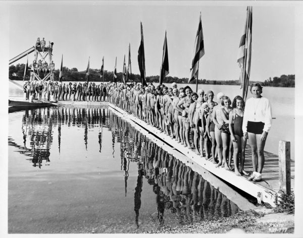 Large group of young women and girls lined up on a pier. A group of them are perched on a diving platform on the end. A man and woman are in a boat in front of the diving platform. Everyone except the coach (right side) is wearing a bathing suit. Eleven flags are spaced along the pier. The wooded shoreline can be seen in the background.