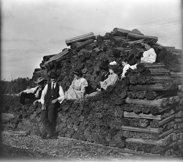 A group of people, consisting of men, women and children, posed on a pile of wooden lath bundles. One man is standing.