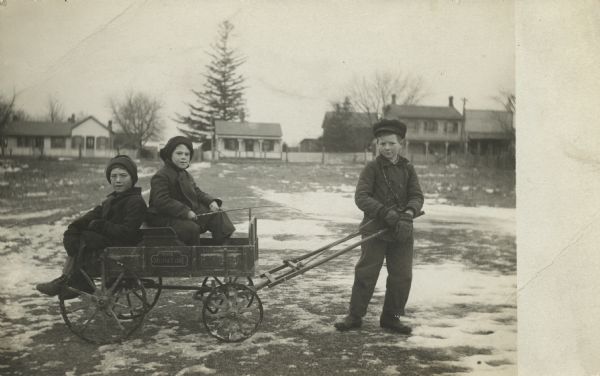 Three boys pose outdoors in a field with snow. Two boys, one holding a stick or a whip, are sitting in a miniature wagon. The other boy is standing holding the wagon tongue. "The Miniature" is on the side of the wagon. Houses, trees and a fence are in the background. The boys are wearing winter clothes.