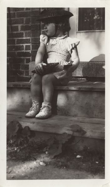 Jo-Claren Maurin (b. 1942) sitting on the stoop of her house in Wauwatosa, Wisconsin wearing a red straw hat and holding a matching red straw purse. She is also wearing a dress, shoes, and socks.