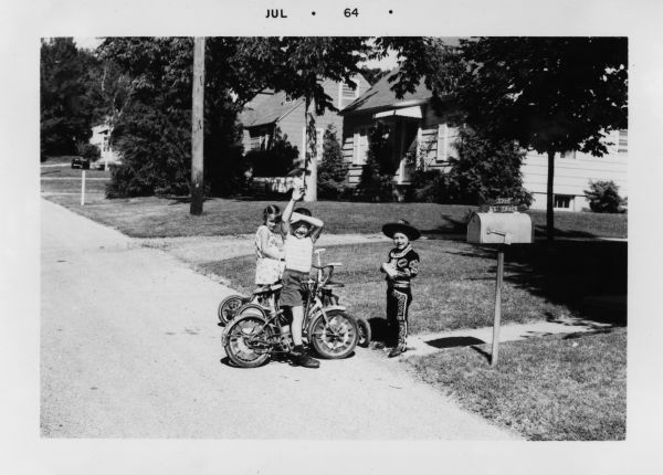 Two children with a tricycle and a bicycle, and one boy in a Mexican cowboy costume, pose at the edge of the street in front of a home, next to a mailbox. There is another tricycle behind the boy who is waving at the photographer. The bikes are decorated for the Fourth of July. Dwellings can be seen in the background.

Michael A. Faber (b. 1957) is the boy waving.  His brother Peter W. Faber (1959-2007) wears a Mexican cowboy costume, originally worn by his father Robert E. Faber (b. 1933) in 1938.  Their neighbor Mary Ann Heasley stands behind Michael.  They are in front of the Faber home at 5706 Dogwood Place, Madison, Wisconsin.