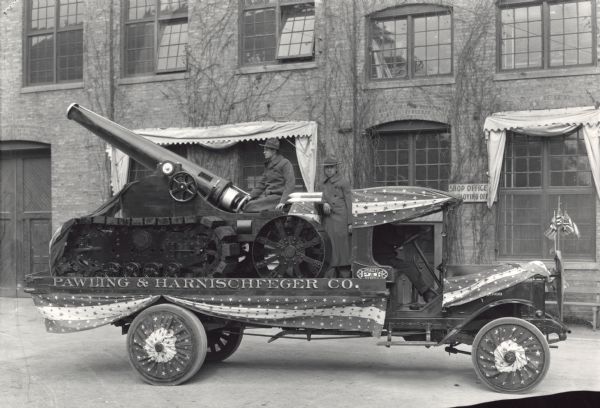 A Pawling and Harnischfeger Company truck, with an artillery gun mounted on the bed, decorated with banners and flags. Two soldiers are standing on the back of the truck. A brick building with many windows is in the background.