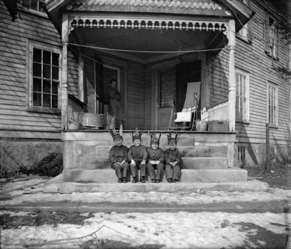 Four boys dressed in Indian costumes sit in a row on the concrete steps to a large porch of a house. A woman wearing an apron is standing on the porch on the left. The porch has two doors leading into the house and has ornate decorations. A washtub, sling chair, clotheslines and other articles can be seen on the porch. There is snow on the sidewalk and in the yard.
