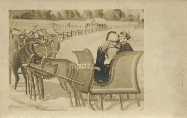 Photographic postcard of Santa and a young girl, Mary Jane Turner, in his sleigh. Her head is next to his and her arm is around his shoulders. Reindeer can be seen on the left and a fence, house and trees are in the background. The image appears to be a photomontage. Except for the two people, the image is painted. The numbers "5671" appear to be glued onto the front of the sleigh and the girl and Santa are tucked inside. The image was then re-photographed and printed on postcard stock.