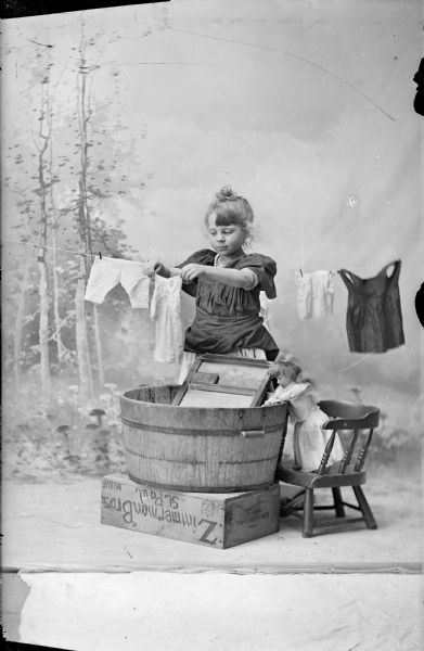 Studio portrait in front of a painted backdrop showing Bertha Gesell washing and hanging doll clothes.