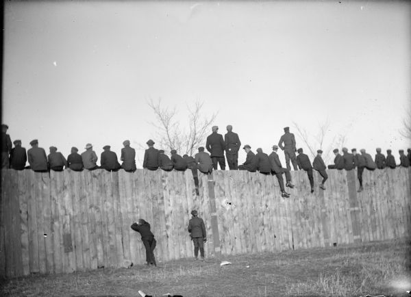 Group watching a University of Wisconsin football game (?) from a fence. Most are sitting on top, a few are standing and two men are peeking through the fence. Graffiti message (difficult to see) written on fence reads, "Millions for defence but not 1¢ for Tribute."

