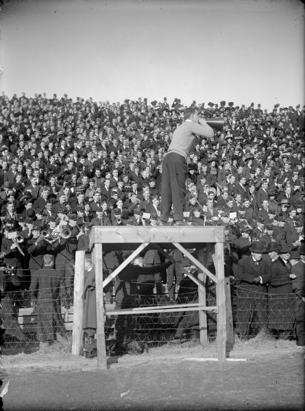 A male cheerleader with megaphone leading the crowd at a University of Wisconsin football (?) game. The band performs at the front of the crowd.