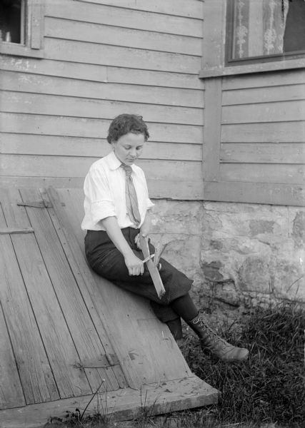Young woman, possibly a University of Wisconsin student, whittling while seated outdoors on a cellar doorway or bulkhead.