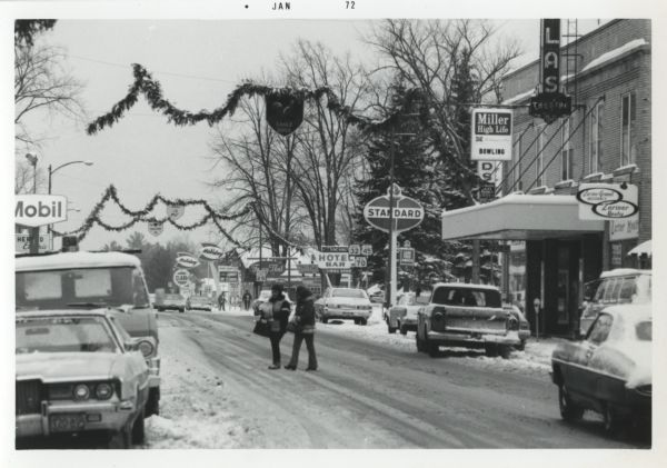 Winter scene on Wall Street. Two women cross the street and many cars are parked at the curb. Men appear in the background. Many business signs can be seen and holiday decorations are still on display overhead.