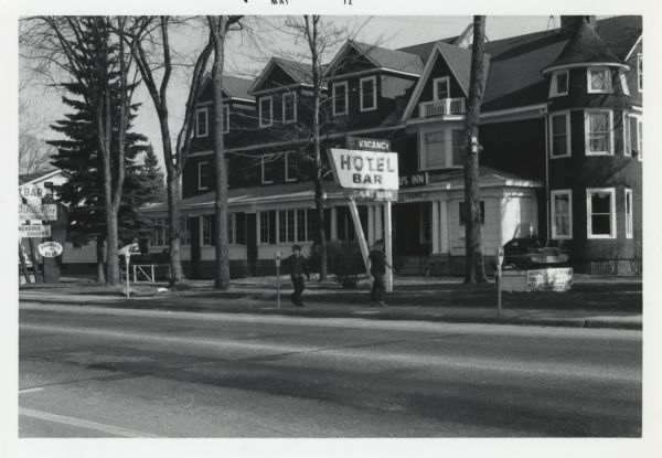 Spring scene on Wall Street. The building is the Arbutus Inn, hotel, bar and dining room. It was also the original Snowmobile Hall of Fame. Two boys are strolling along the sidewalk. On the right is a snowmobile on a pedestal, a sign attached reads, "Eagle River, Snowmobile Capital of the World, Home of the World Championship Snowmobile Derby." Another snowmobile on a pedestal appears on the left.