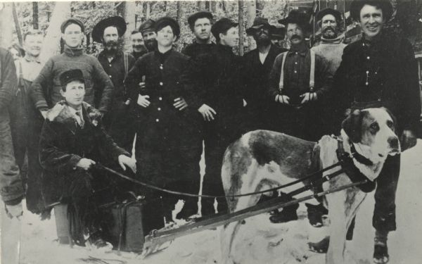 Group portrait of loggers in a lumber camp. In front, seated on his sled, is T.J. Thompson. He traveled among the lumber camps between Winter and Raddison, selling watches and jewelry. His sled was pulled by his dog.