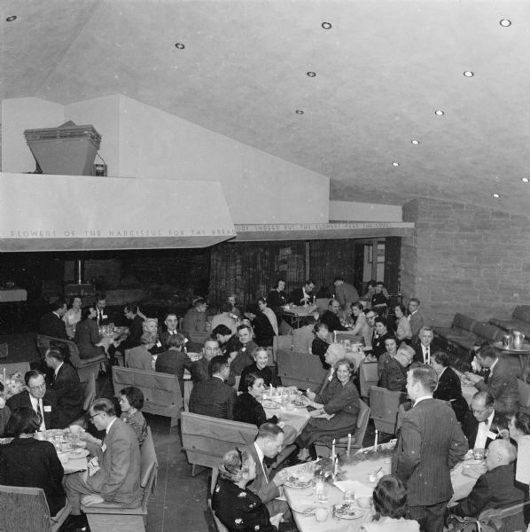 Rev. Max Gaebler, standing at the lower right, addressing a gathering at the First Unitarian Society Meeting House. This view, looking toward the rear of the auditorium, highlights the benches designed by Frank Lloyd Wright to be used for both services and social gatherings. Facing the camera in the middle of the picture is noted economist Arthur Altmeyer, a member of the congregation. At the same table, and facing the left, is William Gorham Rice.