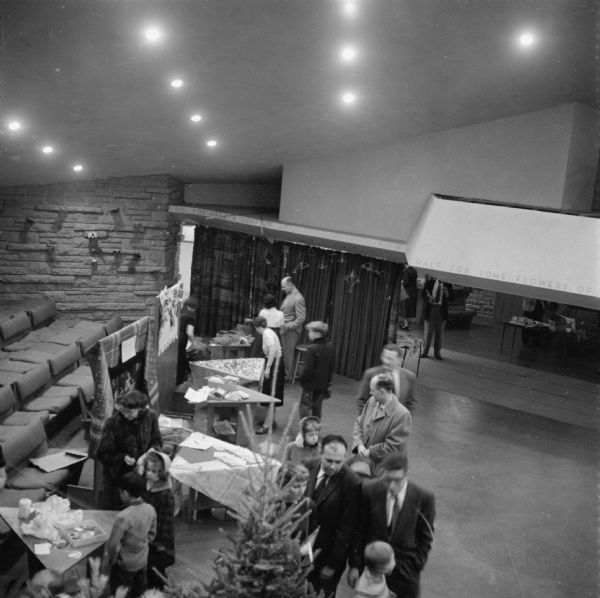 This view of a holiday fund raising event in the auditorium of the Frank Lloyd Wright-designed First Unitarian Society meeting house shows the woven curtain that could be drawn to divide the auditorium into two spaces. The curtain was designed by Wright and woven by members of the society on Taliesin looms. This image also shows the special benches and triangular tables designed by Wright. The top of an evergreen tree is in the foreground.