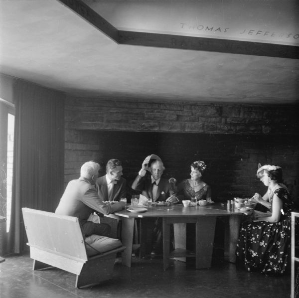 Frank Lloyd Wright in the Hearth Room of the First Unitarian Society Meeting House. Wright was there to give a speech to be aired later on the Omnibus television program. Prior to the speech Wright, the Taliesin Fellows who accompanied him, and members of the First Unitarian Society enjoyed a meal. Olgivanna Wright seated next to the architect had apparently reminded Wright that he should remove his hat. The man next to Wright is thought to be Gerald Bartel, a local broadcasting executive and a member of FUS. The fireplace, which was one of two in the meeting house, is an unusual feature of church architecture.