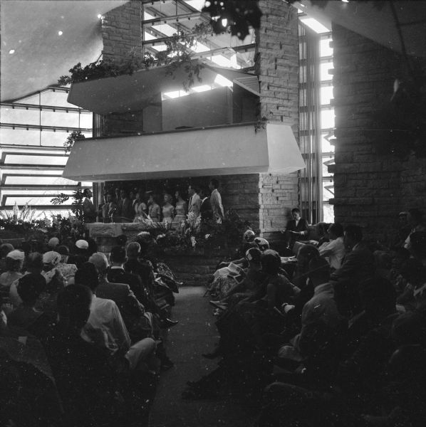 The Taliesin Fellowship choir singing in the pulpit of the First Unitarian Society Meeting House, a Frank Lloyd Wright design. Wright and the Fellowship choir were present to film a segment for the Omnibus television program. Here, the choir performs for the audience who came to hear Wright speak. Normally the First Unitarian Society choir performed in the space above the pulpit. The roof over the choir loft, a tympanum, was designed by Wright to aid the acoustics of the space.