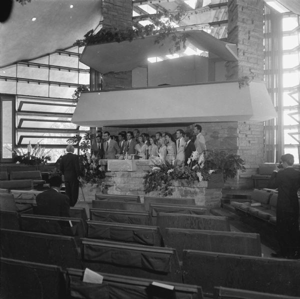 Frank Lloyd Wright visited the First Unitarian Society Meeting House, a building he designed, to be filmed by the Omnibus television program. In addition to a speech by Wright, the filming included vocal selections by the Taliesin Apprentices. In this view of the pulpit they can be seen rehearsing, while Mr. Wright, facing the choir, listens. A photographer snaps a picture on the right.