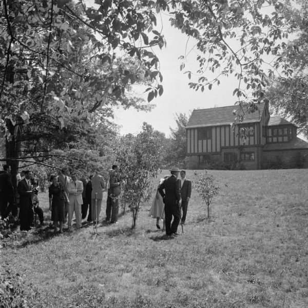 Frank Lloyd Wright (near the center of the image) and friends touring the grounds of the First Unitarian Society meeting house.  In the background is the Tudor Revival Isom house, also on the property, which served as the parsonage. It is assumed that Wright disliked the proximity of the Isom House to the building he designed.