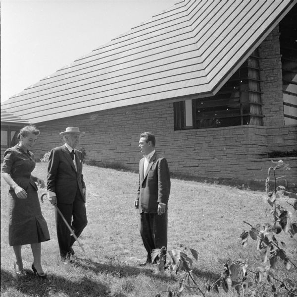 Frank Lloyd Wright touring the grounds of the First Unitarian Society Meeting House, a Wright-designed structure. Behind Wright is the soaring roof of the meeting house auditorium, the Prow, the iconic element of this design. The two people with Wright have not been identified.