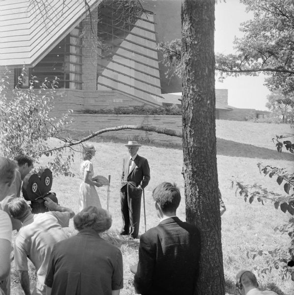 Architect Frank Lloyd Wright being filmed for the Omnibus television program. Wright and apprentice Frances Nemtin are standing in front of the Unitarian Meeting House that Wright designed. Although Wright was interviewed live on the program, filming done at the meeting house did not air.