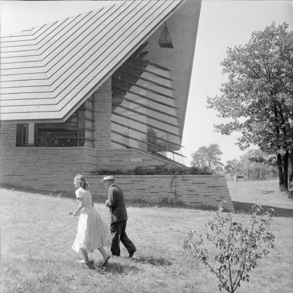 Architect Frank Lloyd Wright touring the grounds of the First Unitarian Society Meeting House, one of his best known works. The woman walking with him is apprentice Frances Nemtin. The iconic roof of the building, the Prow, can be seen behind them. Wright was visiting to be filmed for the Omnibus television program.