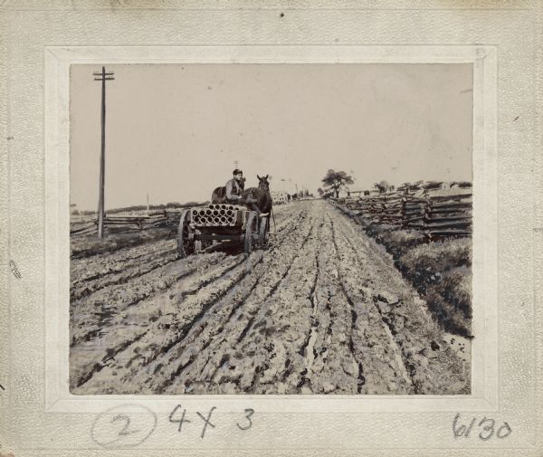 Rear view of a man driving a horse-drawn wagon filled with pipe on a muddy, rutted dirt road. A split-rail fence can be seen on both sides of the road and a telephone pole appears on the left. In the far distance are a farm house, buildings and trees. This image was entered in a 1898 competition sponsored by the League of American Wheelman to identify the nation's worst road conditions. The purpose of the competition was to gather evidence of the need for better roads. The donor, Otto Dorner of Milwaukee, was the national chair of the league's road improvement committee.