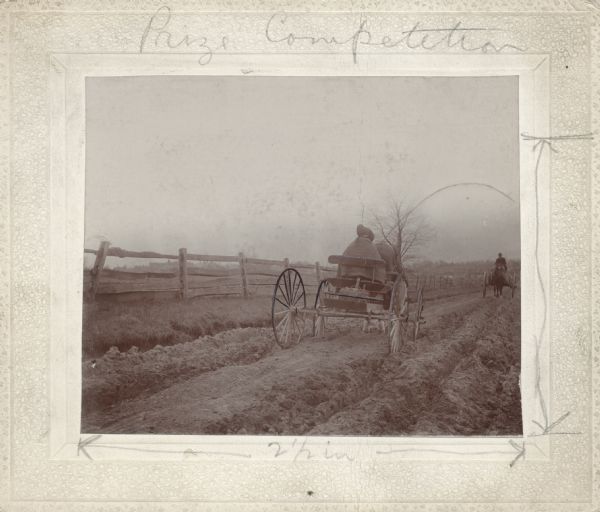 Two horse-drawn wagons travel toward each other on a rutted, dirt road. It appears that a woman wearing a bonnet and shawl is driving the nearer wagon, with a man driving the far wagon. A split-rail fence is on the left. State Normal School buildings can barely be seen on the left and others in the background. This image was entered in a 1898 competition sponsored by the League of American Wheelman to identify the nation's worst road conditions. The purpose of the competition was to gather evidence of the need for better roads. The donor, Otto Dorner of Milwaukee, was the national chair of the league's road improvement committee.