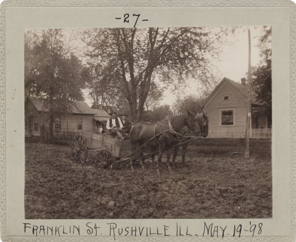 Two men sit in a horse-drawn wagon mired in the mud on a dirt road. Several houses are in the background, with two women standing on a porch on the left, indicate that the road may pass thorough a neighborhood. This image was entered in a 1898 competition sponsored by the League of American Wheelmen to identify the nation's worst roads conditions. The purpose of the competition was to gather evidence of the need for better roads.
