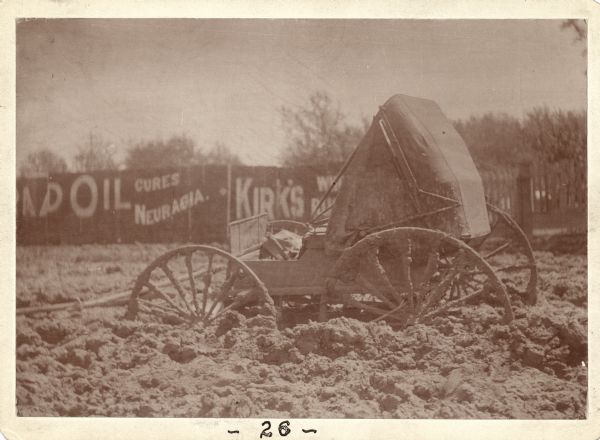 A buggy is mired up to the axles in mud. The horses have been unharnessed and led away. Hand-painted signs are on the fence in the background. This image was entered in a 1898 competition sponsored by the League of American Wheelman to identify the nation's worst roads conditions. The purpose of the competition was to gather evidence of the need for better roads.