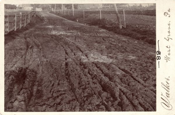 Cabinet card of a deeply rutted dirt road, which leads to buildings in the background. Fences are visible on both sides. This image was entered in a 1898 competition sponsored by the League of American Wheelman to identify the nation's worst roads conditions. The purpose of the competition was to gather evidence of the need for better roads.