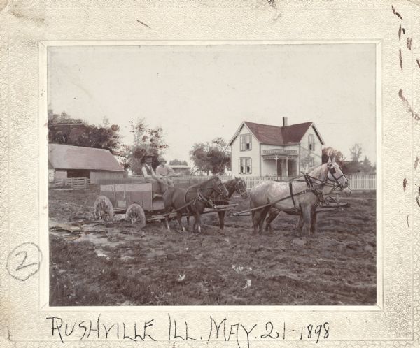 Two men sit in a wagon pulled by four horses, which is stuck in the mud. Farm buildings are on the left and a farmhouse is on the right. This image was entered in a 1898 competition sponsored by the League of American Wheelman to identify the nation's worst roads conditions. The purpose of the competition was to gather evidence of the need for better roads.