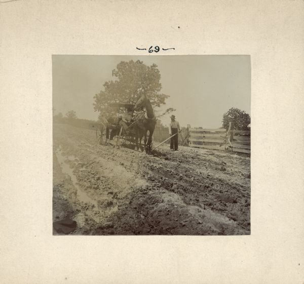 A man sits in a horse-drawn buggy while another man appears to be digging dirt out from under a front wheel. Fences and a gate are on the left. This image was entered in a 1898 competition sponsored by the League of American Wheelman to identify the nation's worst roads conditions. The purpose of the competition was to gather evidence of the need for better roads.
