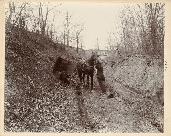 Two men attempt to move a buggy that has tipped over in the mud on a rutted, dirt road with steep embankments on both sides. One man is steadying the horse and his hat is laying in the middle of the road. The other man is under the buggy, trying to push it back up. This is one of a series of photographs collected by the League of American Wheelmen in 1898 from throughout the Midwest in order to publicize the need for better roads.
