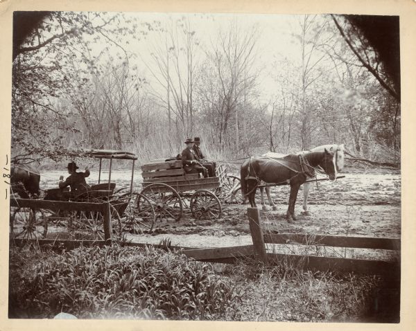 View over fence of two men sitting in their horse-drawn wagon that appears to be stuck in the mud on a rutted dirt road. A buggy has just passed them in the opposite direction. This image was entered in a 1898 competition sponsored by the League of American Wheelman to identify the nation's worst roads conditions. The purpose of the competition was to gather evidence of the need for better roads.