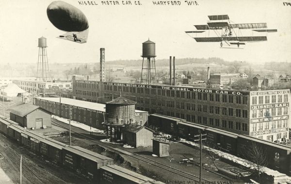 Photographic postcard view of the Kissel Motor Car Company. The elevated view shows the factory buildings, water towers, railroad cars and track. A dirigible and bi-plane appear to have been added to the scene when printed. Caption reads: "Kissel Motor Car Co., Hartford, Wis."